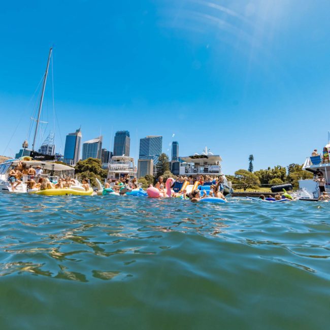 People are floating on inflatable rafts and boats on a sunny day in a city harbor with tall buildings in the background, enjoying the vibrant scene. It’s an ideal day for a private yacht charter Sydney Harbour.