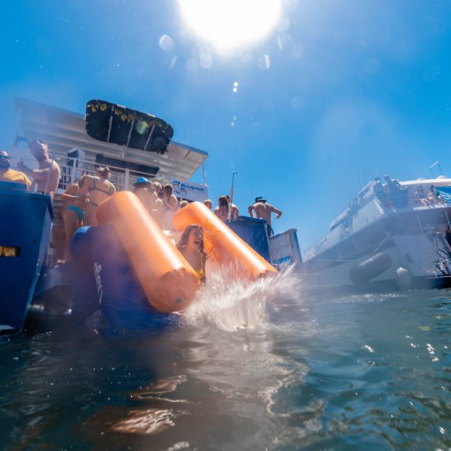 Several people slide into the water from a boat under a bright sun, with another boat visible in the background. It appears to be a lively Sydney boat party hire event.