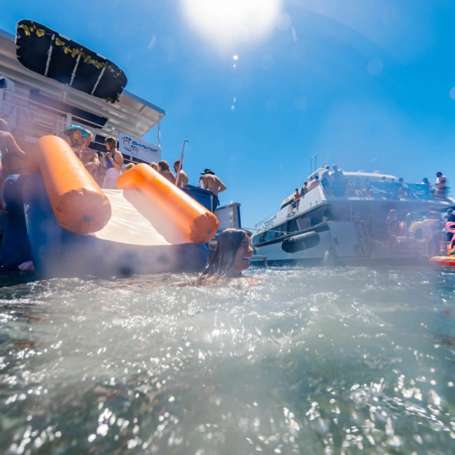 People enjoying a sunny day on boats, some lounging on deck while others swim in the water, surrounded by inflatable floaties and water slides. Ideal for catamaran party Sydney enthusiasts looking to celebrate in style!