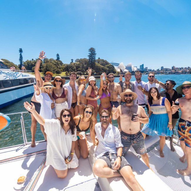 A group of people in swimwear and summer clothing pose and smile for a photo on a private yacht charter in Sydney Harbour, with water, trees, and another boat in the background on a sunny day.
