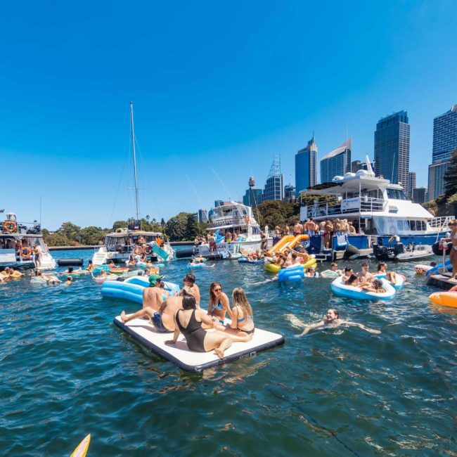People on inflatables and boats enjoying a sunny day on the water with a city skyline in the background, including a luxury yacht hire Sydney experience.