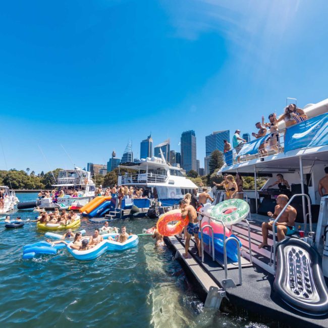 People are gathered on boats and inflatables, enjoying a sunny day on the water with a city skyline in the background. It's the perfect setting for a Sydney boat party hire or a private yacht charter Sydney Harbour.