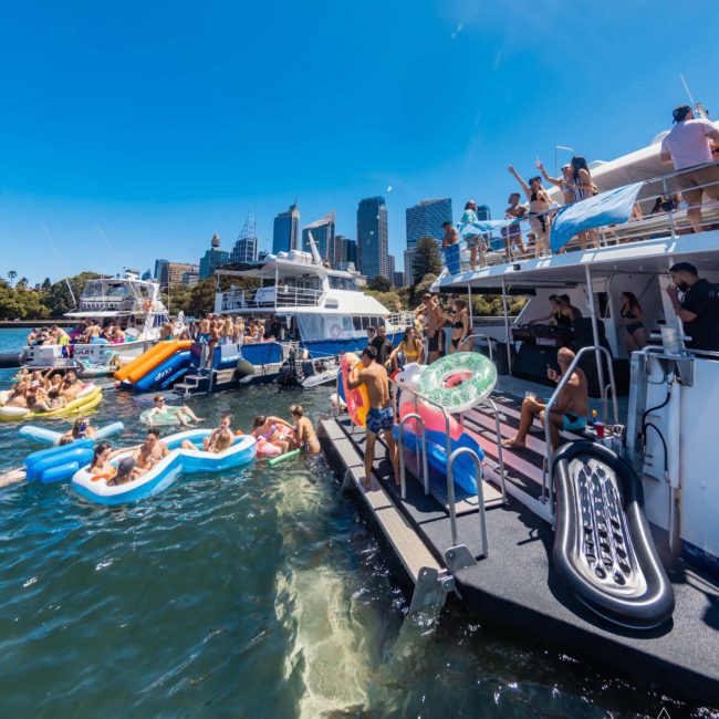 People on various boats and inflatables enjoy a sunny day on the water near a city skyline. The scene includes activities like swimming, lounging, and socializing. A private yacht charter in Sydney Harbour adds an element of luxury to the vibrant atmosphere under the clear blue sky.