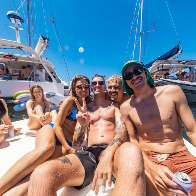 A group of people in swimwear are sitting and smiling on a floating platform in a marina, with boats and more people in the background on a sunny day, enjoying their catamaran party Sydney.
