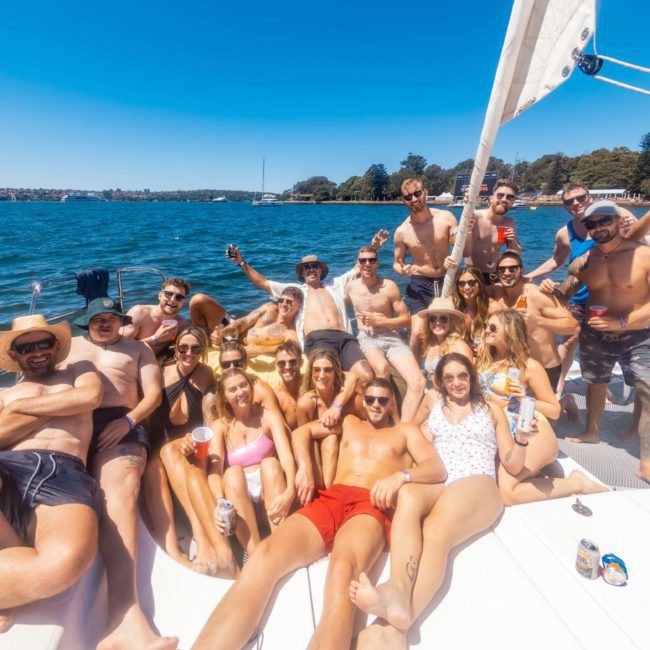 A group of people are gathered on a boat, posing and smiling. Some are sitting, others standing, under a clear blue sky with water and land in the background—perfect for a Catamaran party Sydney.