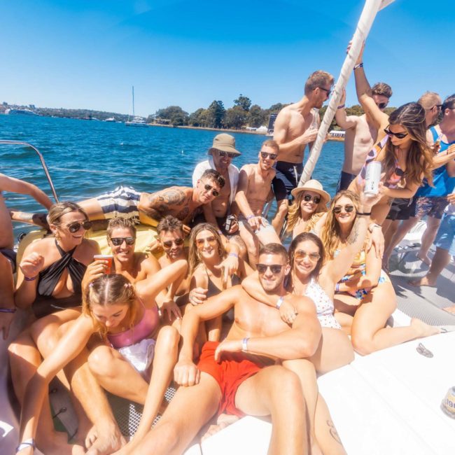 A group of people in swimwear are gathered on a private yacht charter in Sydney Harbour enjoying a sunny day on the water. Some are sitting or standing, holding drinks, while others pose for the camera and bask in the vibrant atmosphere of their corporate boat event.