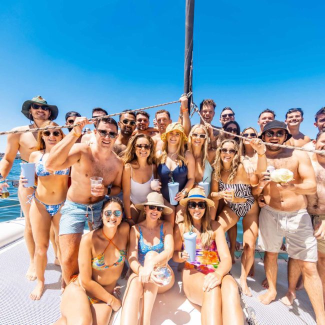 A group of people in swimwear smiling and posing for a photo on a luxury yacht under a clear blue sky, enjoying their time during a Sydney boat party hire.