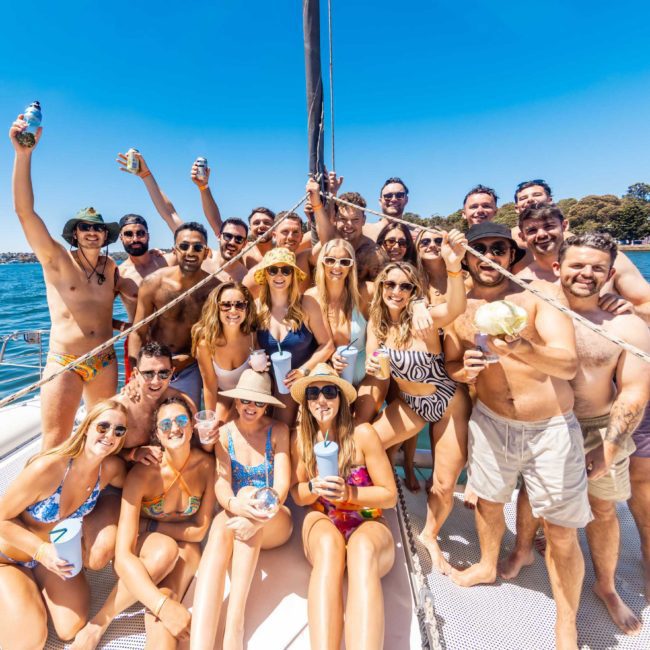 A group of people in swimwear is gathered on a boat, holding drinks and smiling at the camera on a sunny day. The boat is on a body of water with trees visible in the background, epitomizing the perfect Sydney boat party hire experience.