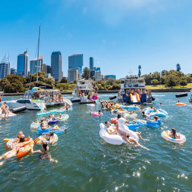 Boats docked near the shore with a city skyline in the background. Numerous people are floating on inflatable devices and swimming in the water on a sunny day, enjoying what feels like a Sydney boat party hire event.