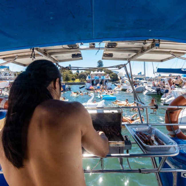Person cooking on a boat's grill while a crowd on other boats and inflatables enjoy a sunny day on the water during a private yacht charter in Sydney Harbour.