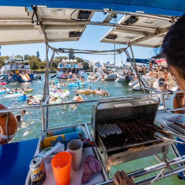 People enjoying a lively catamaran party on a sunny day, with one person grilling food on the boat and others swimming and relaxing on inflatables in the water.