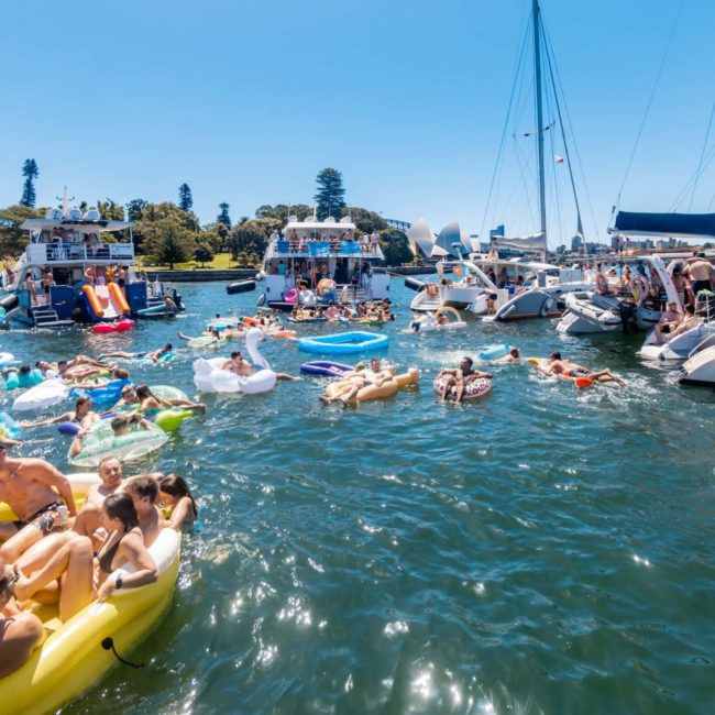 People are gathered on boats and inflatables in a crowded waterway. Some are swimming, while others relax on boats under a clear blue sky. Buildings and trees are visible in the background, making it a perfect scene for a private yacht charter Sydney Harbour or even a lively Sydney boat party hire.