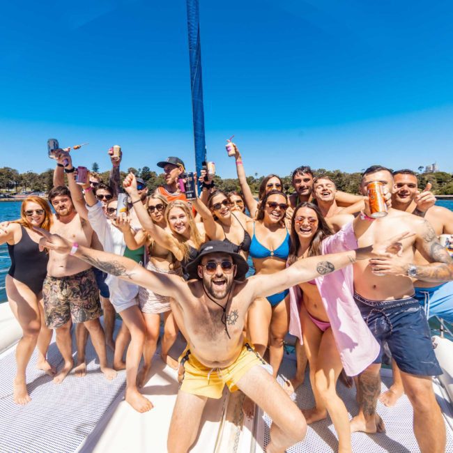 A group of people in swimwear enjoying drinks and posing together on a private yacht charter in Sydney Harbour, with a water body and trees in the background.