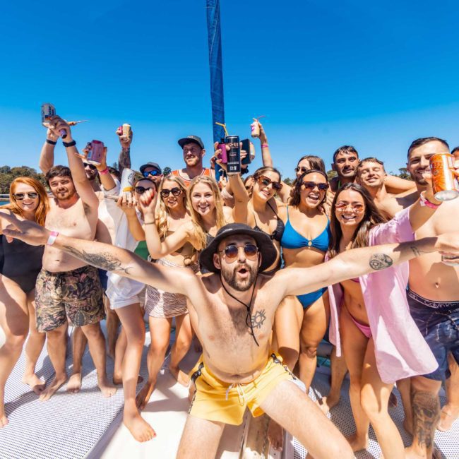 A group of people in swimwear are celebrating on a catamaran party in Sydney under a clear, sunny sky, some holding drinks and wearing sunglasses. Trees and water are visible in the background.