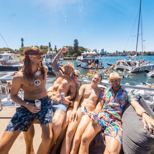 A group of people in swimwear enjoy a sunny day on a private yacht charter in Sydney Harbour, with other boats and individuals visible in the water and background.