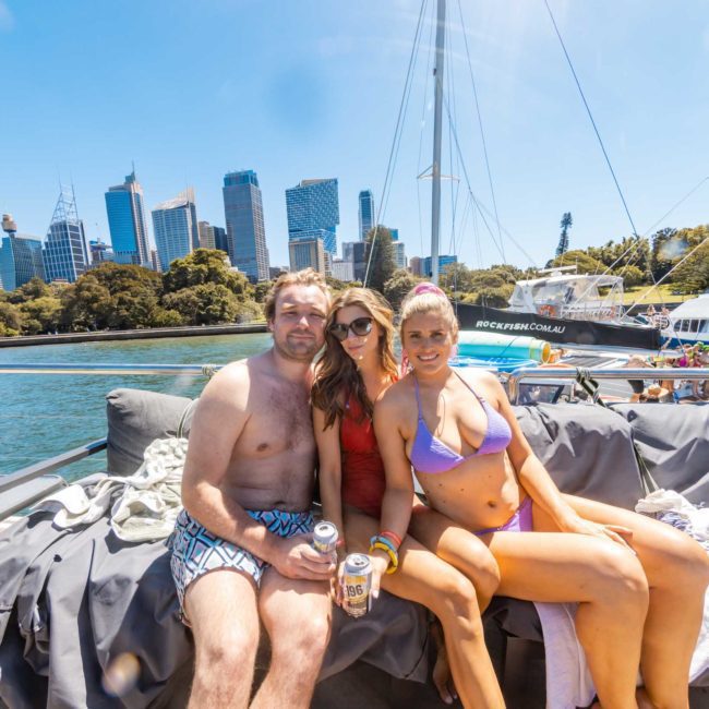 Three people sitting on a private yacht charter Sydney Harbour, with the city skyline in the background. Two women in swimsuits and a man in swim trunks are holding drinks, smiling at the camera.