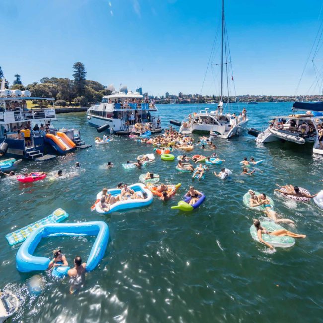 People enjoying a sunny day on a lake, swimming and floating on various inflatables, surrounded by anchored boats. Consider elevating the experience with a DJ boat hire Sydney for your next event.