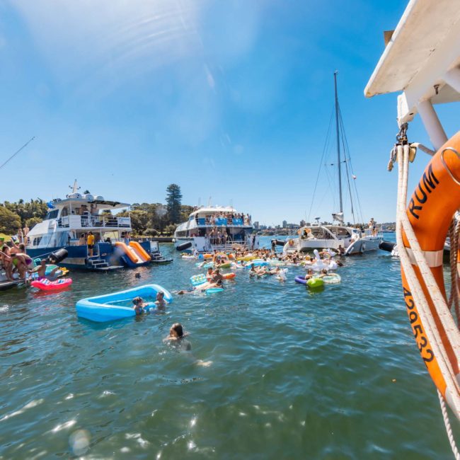 Boats and people with inflatables gather on a sunny day in a body of water. Some individuals swim while others relax on floating devices. An orange lifebuoy is fixed on a nearby boat, highlighting the safety measures often seen in corporate boat events Sydney.