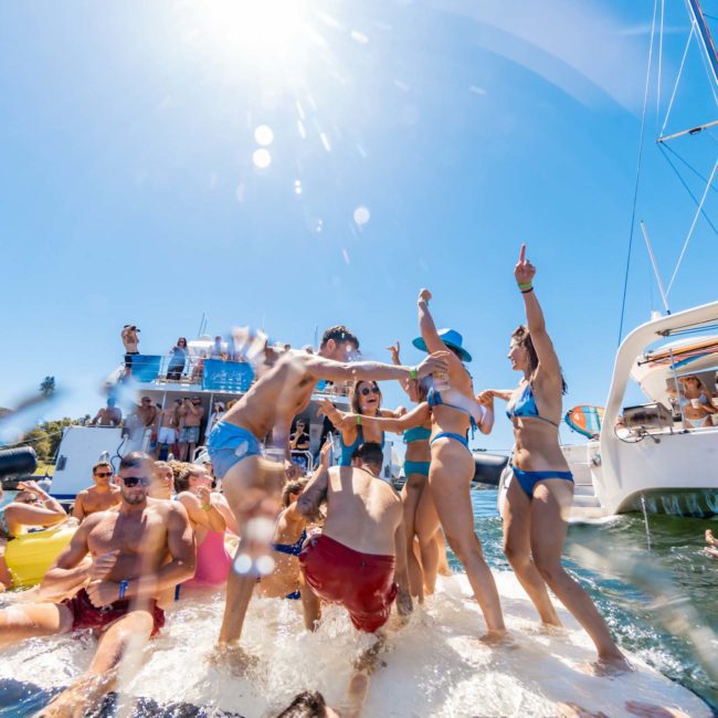 A group of people in swimwear enjoying a sunny day on the water with boats in the background, some dancing and splashing water. The scene looks like a perfect Sydney boat party hire adventure.