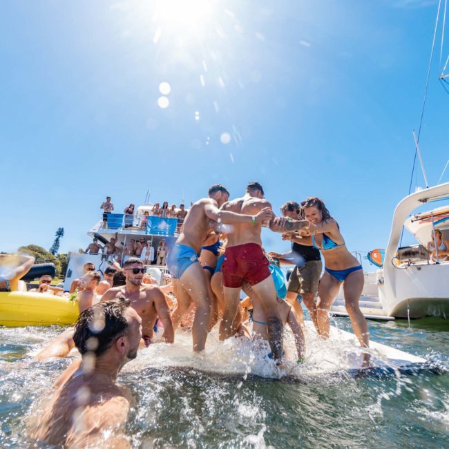 A group of people in swimsuits enjoys a bright sunny day on a crowded catamaran and in the water, some standing on a floating mat, with yachts and city buildings in the background. Perfect for those looking into Sydney boat party hire.