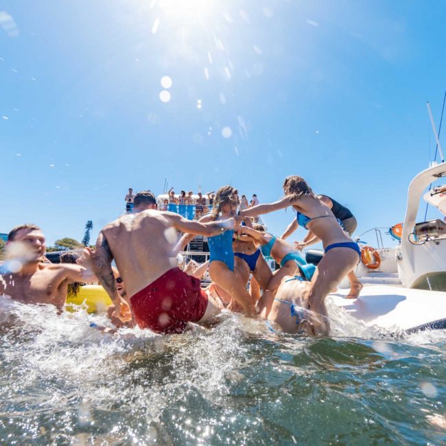 People in swimwear are enjoying a sunny day in and around the water near boats. Some are on inflatable floats and a paddleboard. A luxury yacht hire Sydney adds an elegant touch, with buildings and clear blue sky in the background.