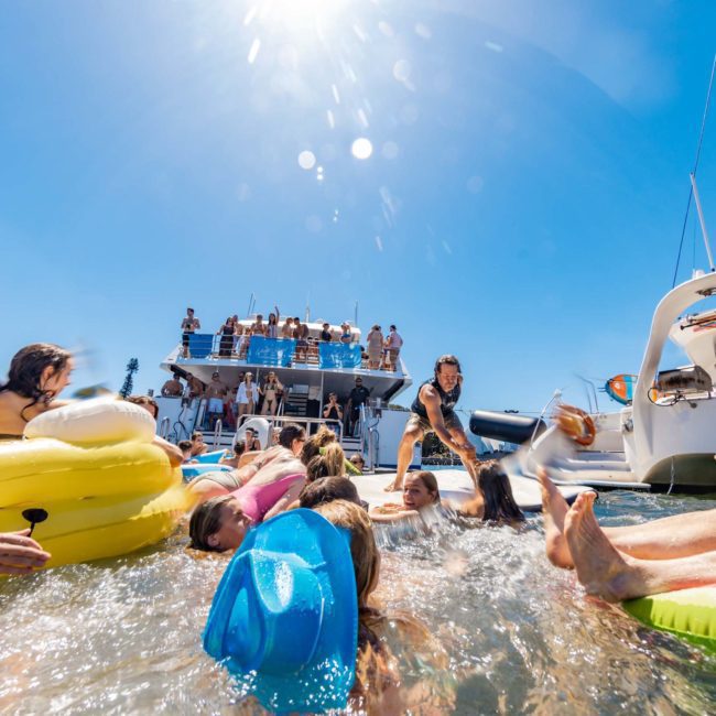 A group of people enjoys a sunny day on a yacht and in the water, using inflatable floats and interacting playfully. The blue sky and Sydney Harbour skyline are visible in the background, highlighting the perfect setting for private yacht charters or corporate boat events.
