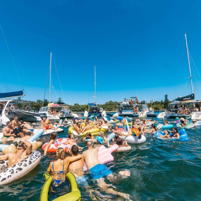 People are gathered on inflatable floats and boats in the water, enjoying a sunny day. Sailboats, including luxury yachts and motorboats, are anchored nearby under a clear blue sky as part of a lively corporate boat event in Sydney.