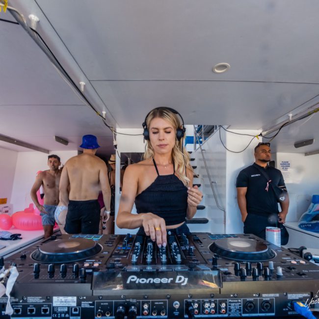 A woman DJ wearing headphones is mixing music on a Pioneer DJ turntable at a private yacht charter in Sydney Harbour. There are several people in the background, some shirtless, standing and talking.