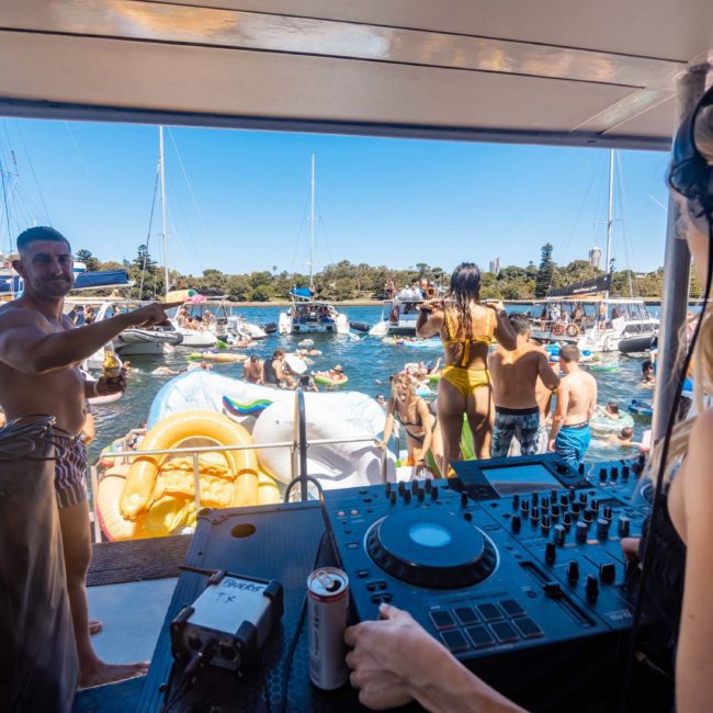 People enjoying a private yacht charter on Sydney Harbour with a DJ playing music, some swimming and relaxing on inflatables, while surrounded by other boats in a sunny location.