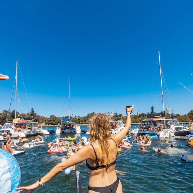 People enjoy a sunny day on the water, surrounded by boats. A person in the foreground raises a drink, while others swim and socialize. The sky is clear and blue, perfect for a private yacht charter Sydney Harbour.