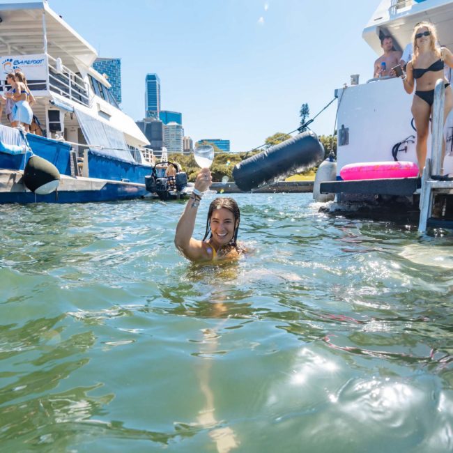 A person swims near a boat in sunny weather, surrounded by other boats and people enjoying the water. The skyline of Sydney is visible in the background, making it a perfect setting for corporate boat events or a lively Sydney boat party hire.