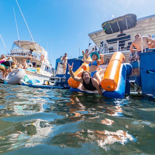 People enjoying a sunny day on boats and inflatables in the water, with a woman sliding and splashing into the water from an orange float—a perfect scene for a private yacht charter Sydney Harbour.