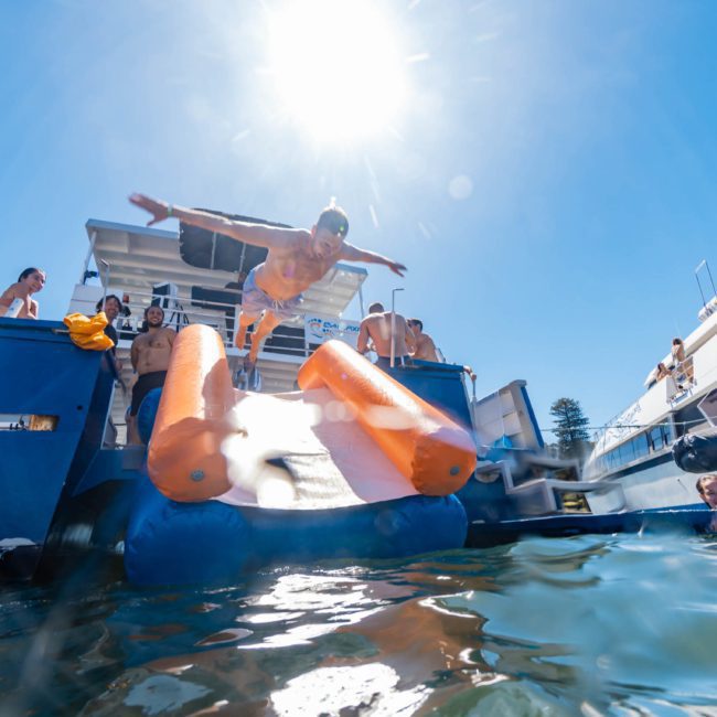 A person in mid-air is diving from a luxury yacht with an orange inflatable slide into the water, surrounded by others on nearby boats under a bright sun. Perfect for corporate boat events in Sydney.