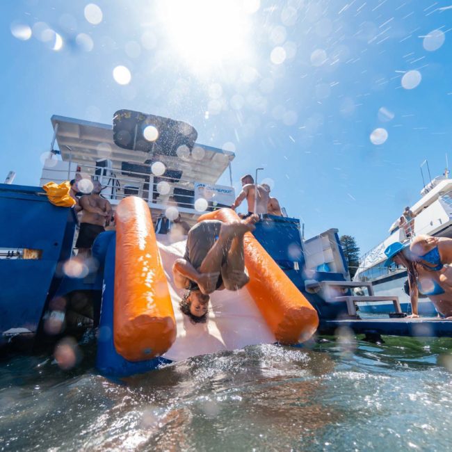 People enjoying a sunny day on a catamaran party in Sydney, with one person about to enter the water. Several others are on the boat and in the water, making the most of this memorable corporate boat event.