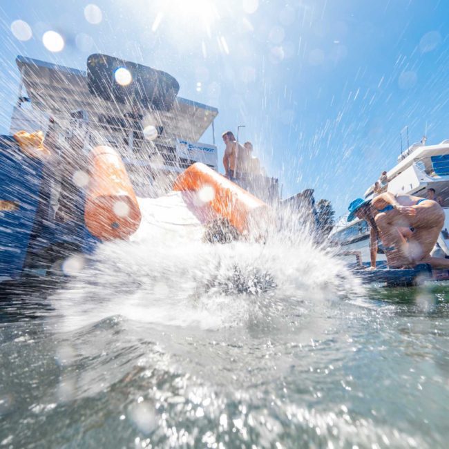 People are enjoying a sunny day on boats, with some using a slide to splash into the water. The image captures a large splash mid-air as someone slides into the water. Consider elevating your experience with Luxury yacht hire Sydney for an unforgettable day out on the water.