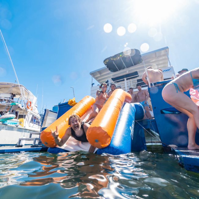 People enjoying a sunny day on and around boats, with several engaging in water activities and using inflatable floats, during a Catamaran party in Sydney.