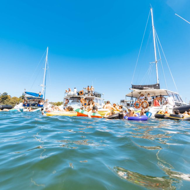 Boats with people on and around them float on a sunny day in clear, blue water, with a bright blue sky overhead. Experience the ultimate scene through a private yacht charter Sydney Harbour has to offer.