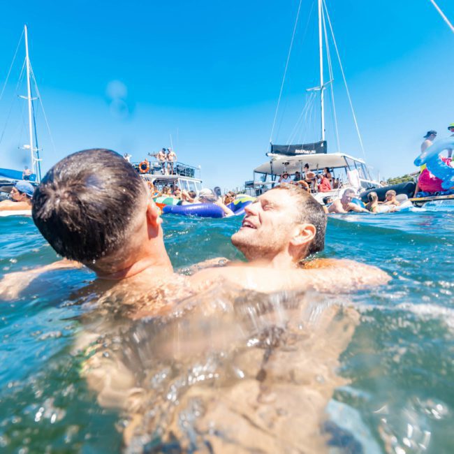 Two people are in the water at a lively Sydney boat party hire, with several boats and other participants visible in the background on a sunny day.
