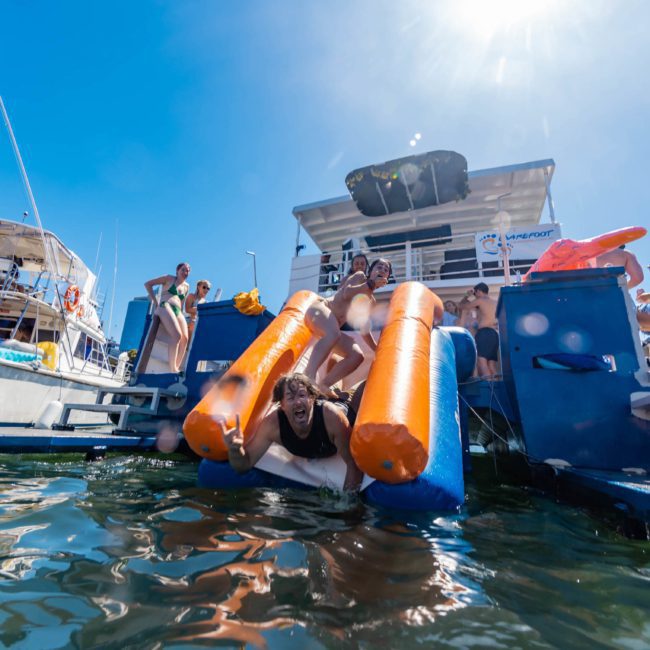 A person slides down an inflatable slide from a private yacht charter in Sydney Harbour into the water, while others stand or sit on the boat in the background under a sunny, clear sky.
