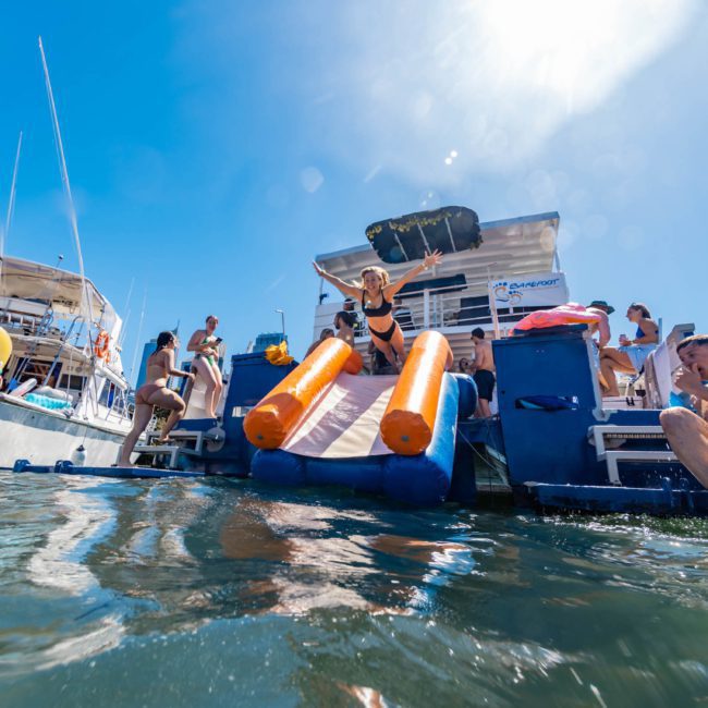 A group of people enjoy a sunny day on a catamaran party in Sydney, with water slides and inflatables, some swimming in the clear water.
