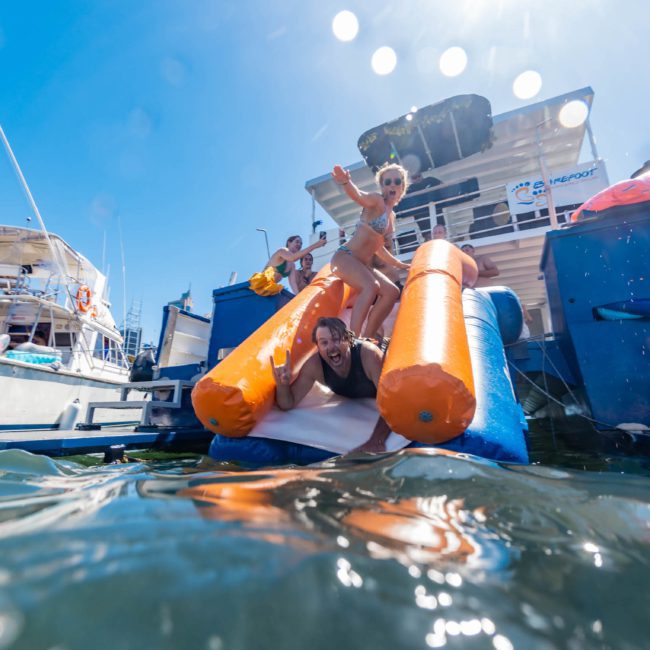 A woman slides into the water from an inflatable slide attached to a luxury yacht, while people watch and enjoy the sunny day on surrounding boats.