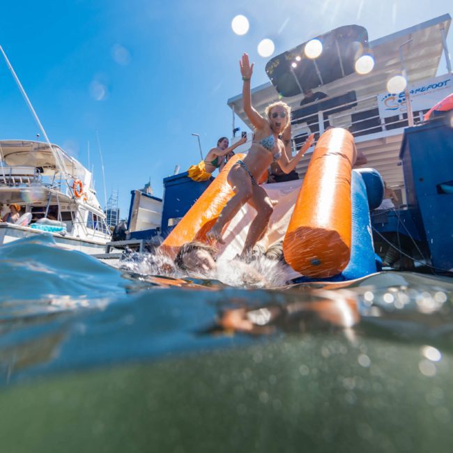 A child wearing swimsuits is sliding down an orange inflatable water slide into a body of water, with boats and people in the background under a bright blue sky, reminiscent of a fun-filled Catamaran party in Sydney.