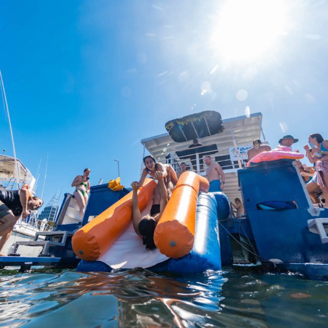People enjoying a sunny day on boats; one person slides down an inflatable slide into the water. Others are standing and sitting on the boats, making the most of their Catamaran party in Sydney.
