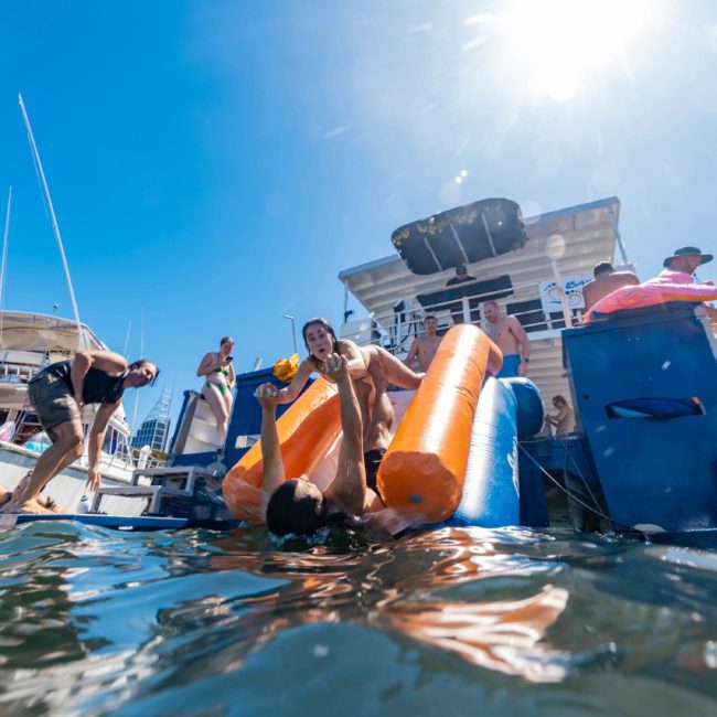 People on boats enjoying a sunny day, some entering the water using orange flotation devices, with a clear blue sky above. Experience the ultimate catamaran party in Sydney for unforgettable moments.
