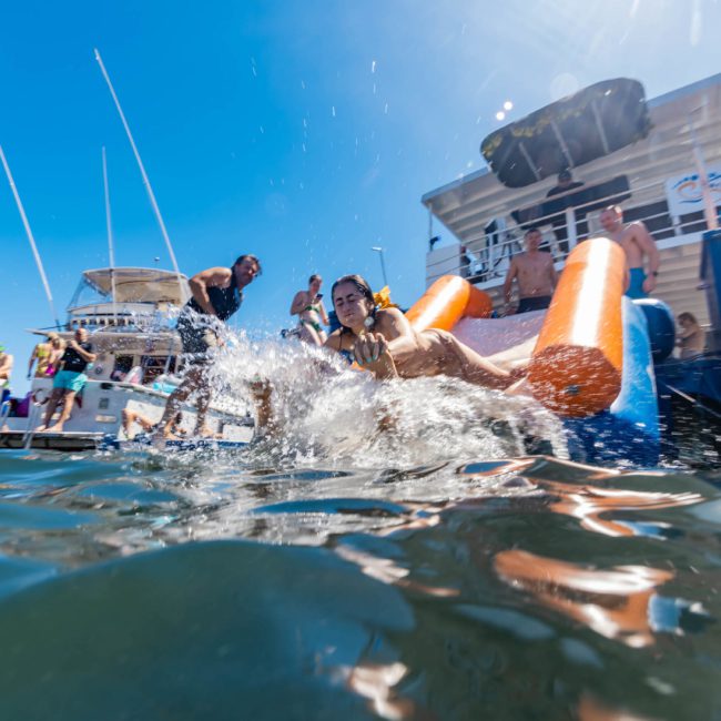 People are enjoying a sunny day on boats docked together. A person is sliding into the water from an orange and yellow inflatable slide, creating a splash. It's a perfect scene for DJ boat hire Sydney or even a vibrant catamaran party Sydney.