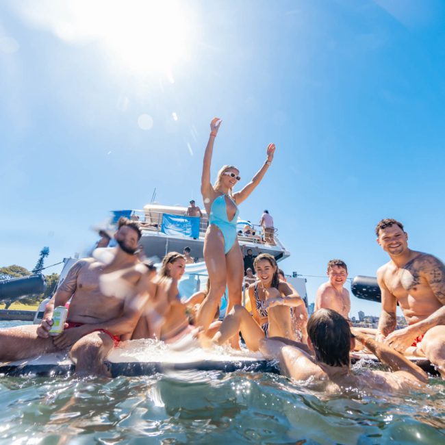 A group of people enjoying a sunny day on a boat, with a woman at the center standing and raising her arms, as part of an exhilarating private yacht charter on Sydney Harbour.