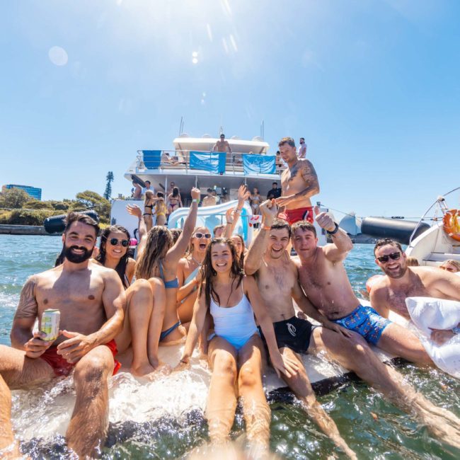 A group of people in swimsuits are sitting and standing on a floating platform in the water, with boats and a cityscape in the background, enjoying a sunny day during their luxury yacht hire Sydney adventure.