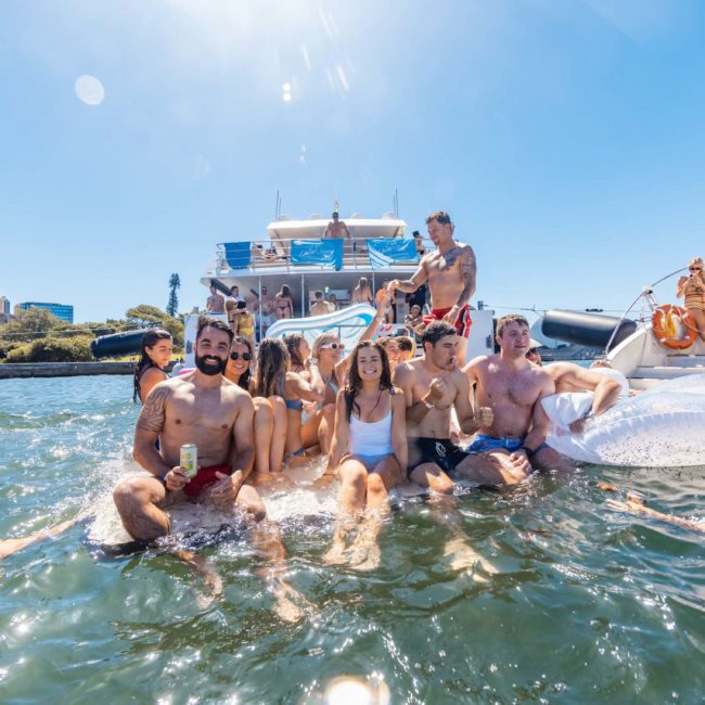 A group of people in swimsuits are enjoying a sunny day in the water, surrounded by inflatable floats and boats, with city buildings in the background. It looks like a scene from an exclusive private yacht charter on Sydney Harbour.