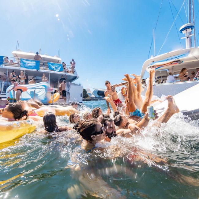 A group of people enjoying a sunny day on a luxury yacht hire Sydney, swimming, sliding, and lounging on floaties, with more guests on the yacht's deck.