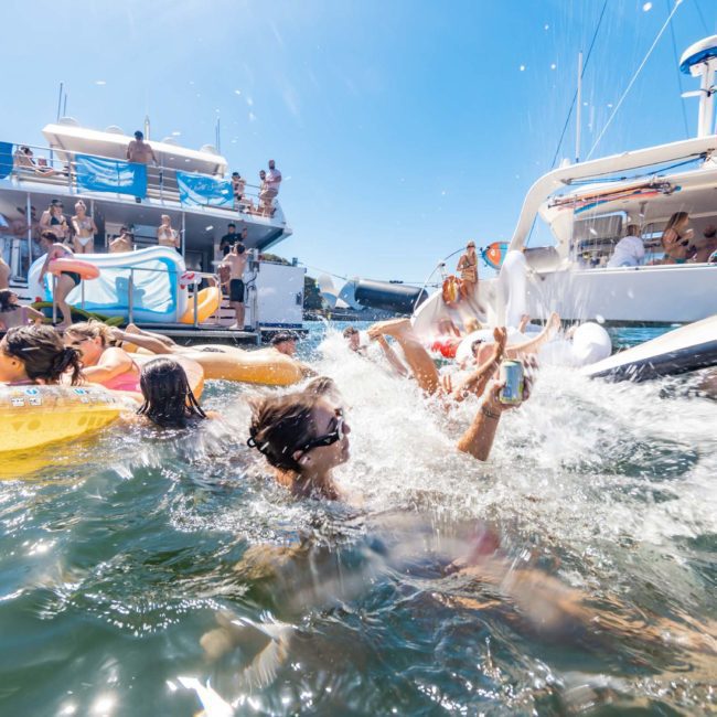 People enjoying a sunny day on the water with boats in the background; some are swimming while others relax on floats, making the most of their Private yacht charter Sydney Harbour experience.